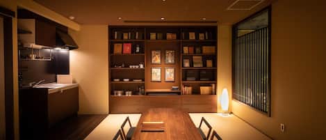 A dining area with a wooden table and chairs, a kitchen with a stove and cabinets, and a bookshelf with various items.