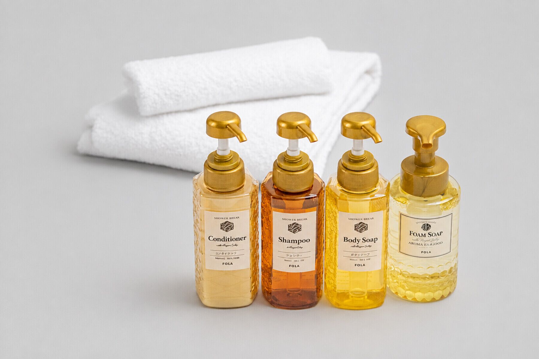 Four bottles of shower products with gold pumps, labeled as conditioner, shampoo, body soap, and foam soap, with a white towel in the background.