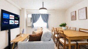 Uma sala de estar moderna com televisão de tela plana exibindo serviços de streaming, uma área de jantar com mesa e cadeiras de madeira, e um ventilador de teto.