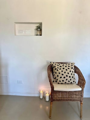 Una silla de mimbre con un cojín de lunares, un aplique de pared y dos velas encendidas sobre el suelo.