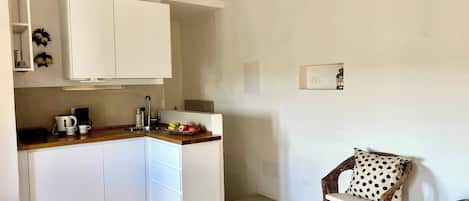 A compact kitchen with white cabinets, a wooden countertop, and a wicker chair with a polka dot cushion.
