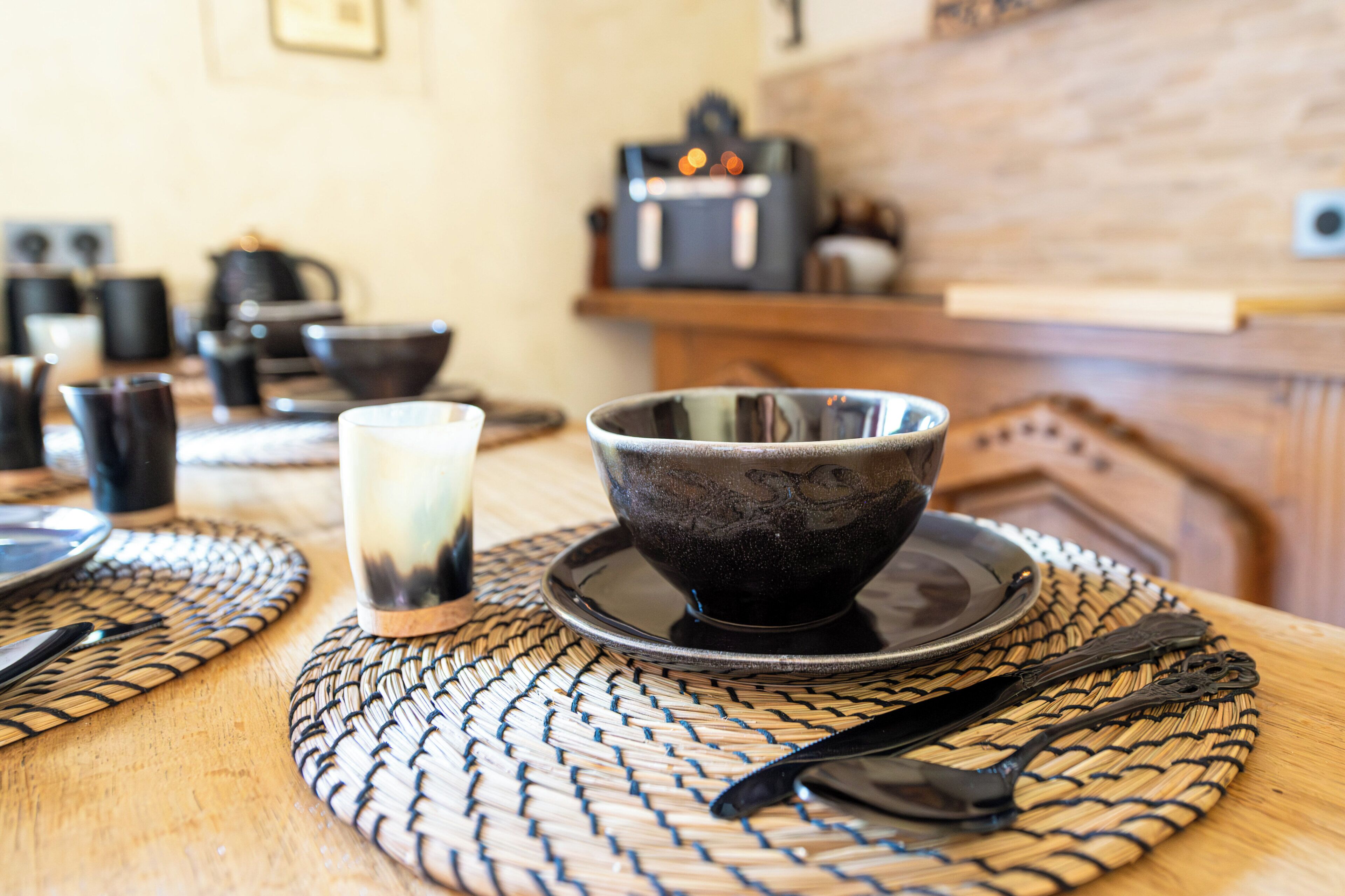 A table with a woven placemat, a black bowl, a glass, and a spoon.