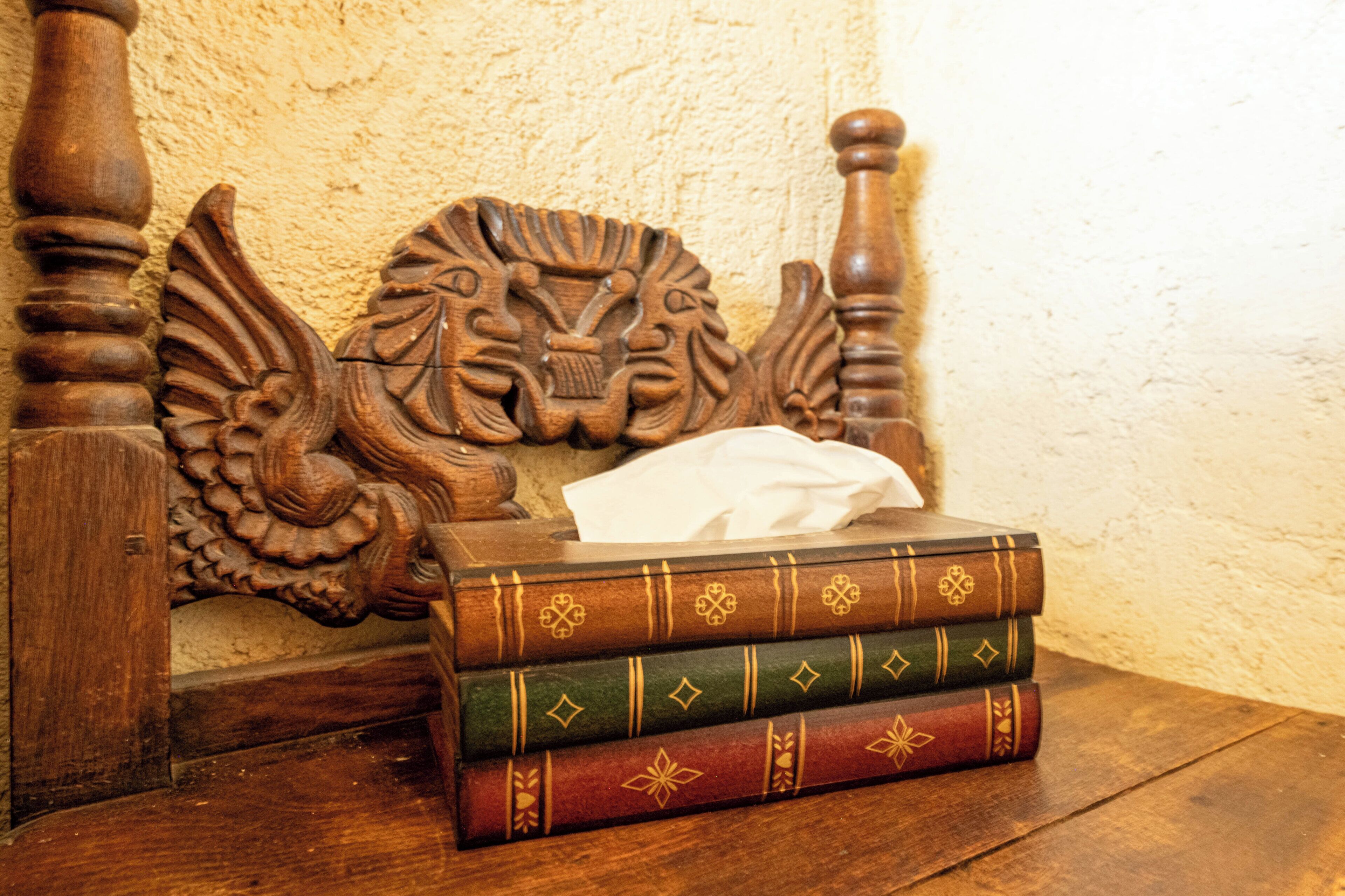 Un lit en bois avec une tête de lit sculptée, une pile de livres et une boîte de mouchoirs posés sur un plancher en bois.