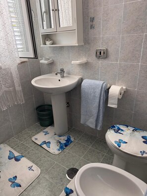 Une salle de bain équipée d’un lavabo, de toilettes et d’un bidet. On y trouve une fenêtre avec des stores, un porte-serviettes et une poubelle.