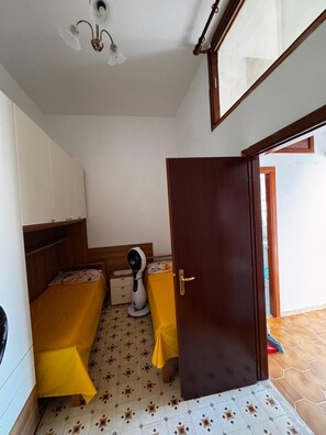 Ein kleiner, kompakter Raum mit einem Bett, einem Nachttisch, einem Ventilator und einer Tür zu einem Außenbereich.