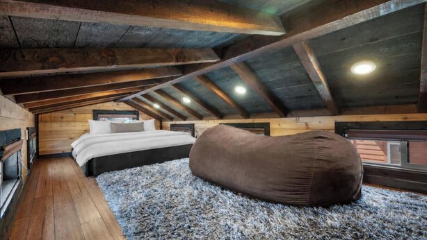 A loft bedroom with a sloped ceiling, a bed, a bean bag, and a view through a window.