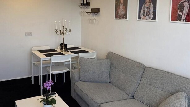 A modern living room with a grey sofa, a white coffee table, a dining table with chairs, a chandelier, and wall-mounted wine bottles. - Apartment Sleeps 2 With Free Parking (Orpington)