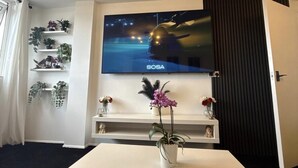 A modern living room with a flat-screen TV mounted on the wall, a white table with a potted plant, and a black carpet. - Apartment Sleeps 2 With Free Parking (Orpington)