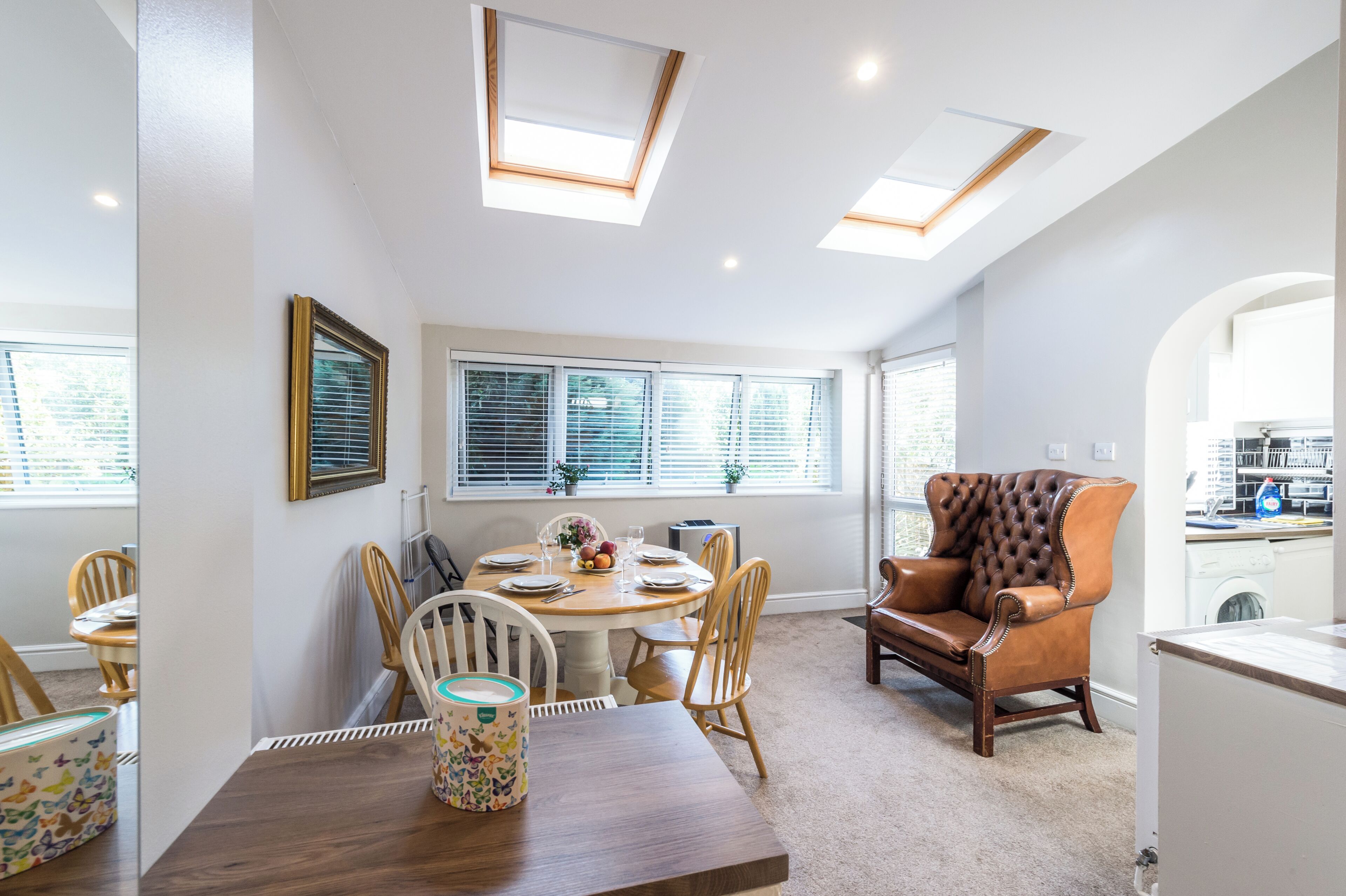 A dining area with a round table, chairs, and a sofa. There are skylights above the table and a window with blinds.