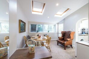 Ein Essbereich mit einem runden Tisch, Stühlen und einer Couch. Über dem Tisch befinden sich Oberlichter und ein Fenster mit Jalousien.