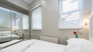 Une chambre d’hôtel avec un lit, un radiateur, une fenêtre avec des stores, un miroir et un vase avec une fleur.