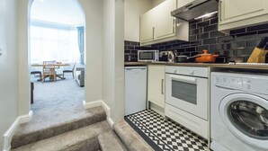 Une cuisine compacte équipée d’un four à micro-ondes, d’un four et d’un lave-linge. Le sol est carrelé et, à travers une arche, on aperçoit un coin repas.