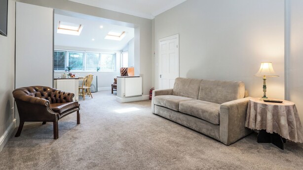 Un salon avec un fauteuil en cuir marron, un canapé beige et une petite table sur laquelle se trouve une lampe. Au fond, on aperçoit un coin cuisine avec une porte blanche et une fenêtre équipée de stores.