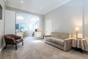 A living room with a brown leather armchair, a beige sofa, and a small table with a lamp. There is a kitchen area in the background with a white door and a window with blinds. - Beautiful Apartment,accomodates up to 6 people,12 minutes tube to Bond street! (London)