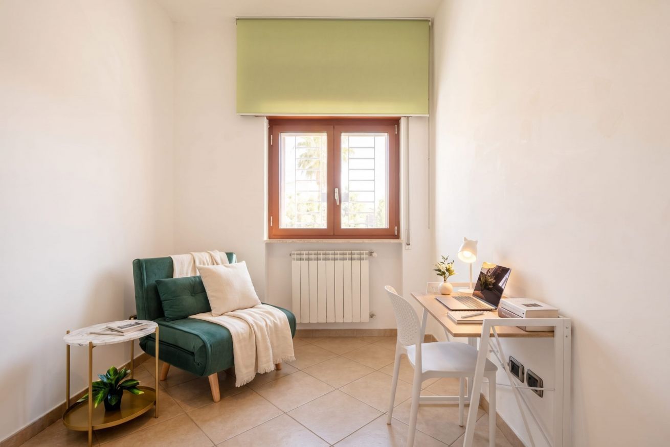 Una stanza piccola e luminosa con un divano verde, un tavolino rotondo con una pianta, una scrivania bianca con un portatile e un termosifone sotto una finestra.