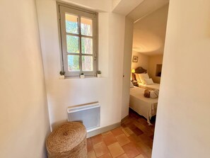 A narrow hallway with a radiator, a window with a view of greenery, and a bed with a headboard and pillows. - Le Bastidon (La Garde-Freinet)