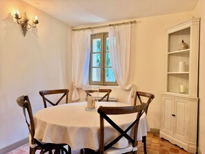 Uma área de refeições com uma mesa redonda, cadeiras de madeira, toalha de mesa branca, uma vela e um armário branco com prateleiras.