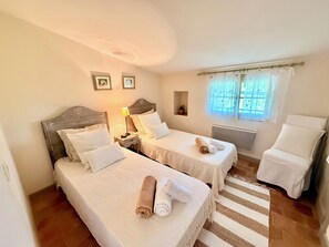 A hotel room with two beds, a chair, a nightstand, a lamp, a window with curtains, and a small shelf with a plant. - Le Bastidon (La Garde-Freinet)