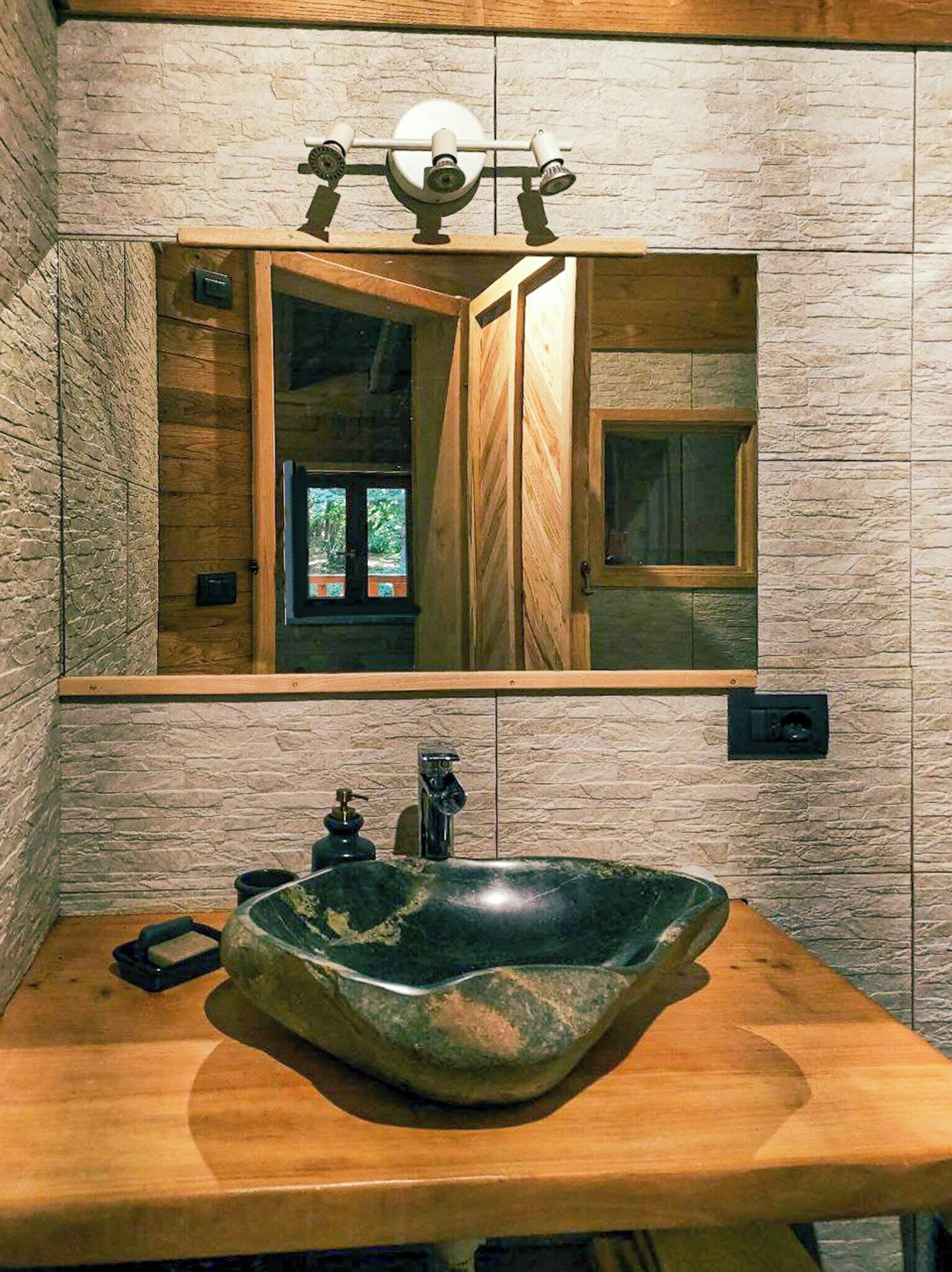 A bathroom with a stone wall, a wooden vanity, a large mirror, and a greenish-brown sink.