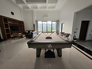 Ein Billardtisch in einem modernen Raum mit einem großen Fenster, einem Fernseher und einer Sitzecke.