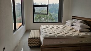 Ein Schlafzimmer mit großem Fenster, einem Bett mit Kissen, einer Bank und Blick auf eine bewaldete Landschaft.