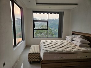 Ein Schlafzimmer mit großem Fenster, einem Bett mit Kissen, einer Bank und Blick auf eine bewaldete Landschaft.