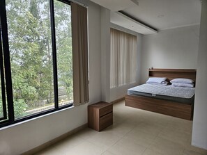 Ein Schlafzimmer mit einem Bett, einem Nachttisch, einem Fenster mit Vorhängen und Blick auf Bäume.