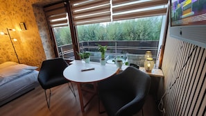 A room with a bed, a table with chairs, a window with blinds, and a potted plant.