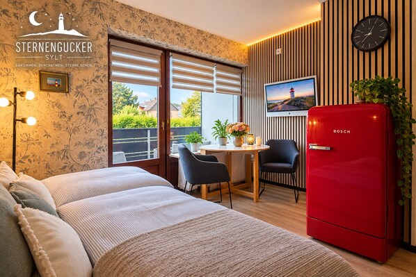A modern bedroom with a bed, a red Bosch refrigerator, a dining table with chairs, and a TV on the wall.