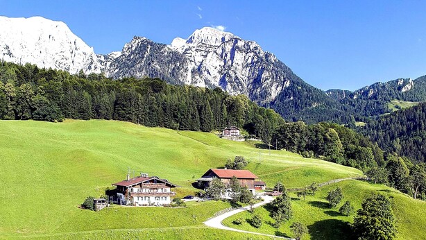 Paisagem montanhosa com casas numa colina verde, uma estrada sinuosa e picos nevados ao fundo.