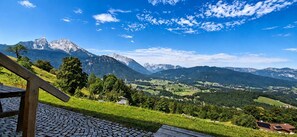 A mountainous landscape with a clear sky, a paved area with a wooden bench, and distant buildings.