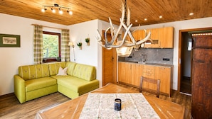Uma sala de estar aconchegante com um sofá verde, uma mesa de jantar de madeira com toalha de mesa estampada e uma área de cozinha com armários de madeira e luminária pendente.