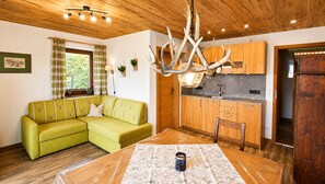 Ein gemütliches Wohnzimmer mit einem grünen Sofa, einem Holzespeisetisch mit gemustertem Tischläufer und einer Küchenzeile mit Holzschränken und einer Pendelleuchte.