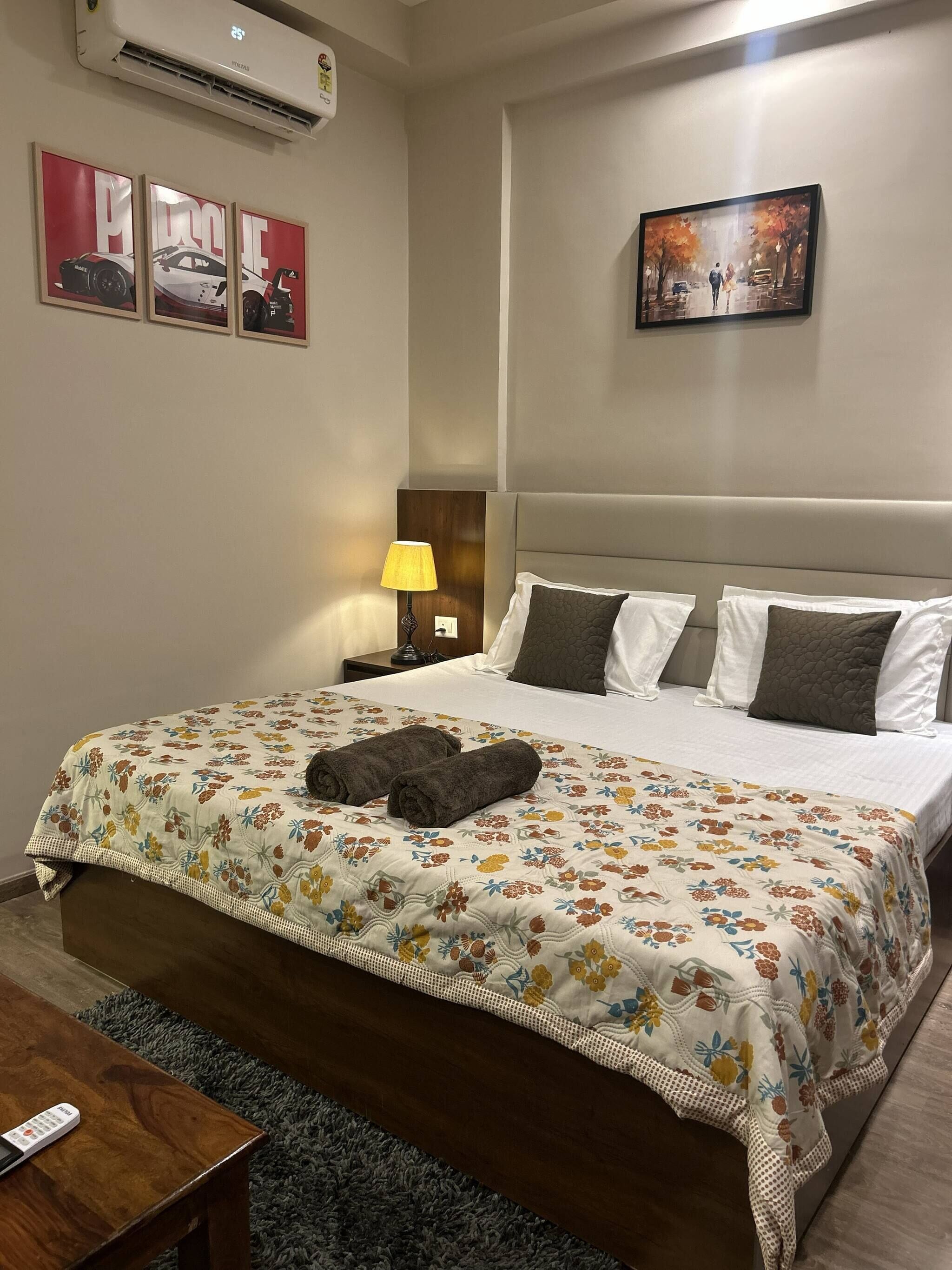 A hotel room with a bed, a nightstand with a lamp, a remote control, and wall art.