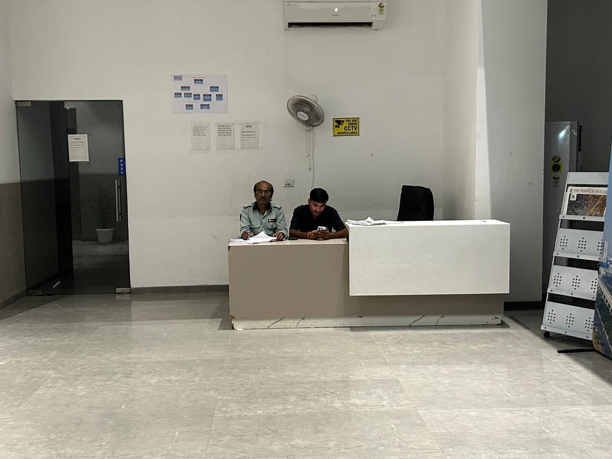 Uma área de recepção com balcão, duas pessoas sentadas, um ventilador fixado na parede e um aviso de circuito fechado de televisão.