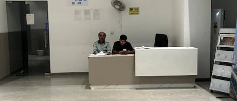 A reception area with a desk, two seated individuals, a wall-mounted fan, and a CCTV sign.