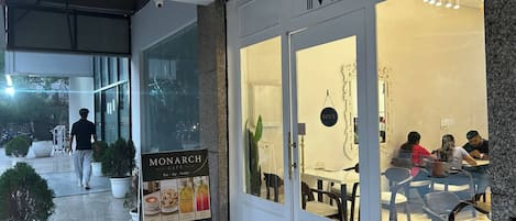 An outdoor seating area of a café with a glass wall, a signboard advertising the café's offerings, and a few people dining inside.