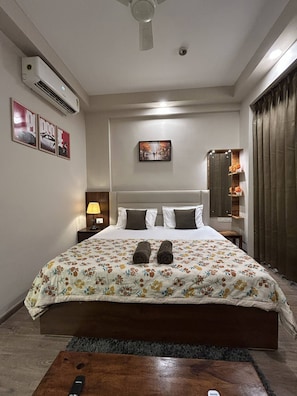 A hotel room with a bed, a nightstand, a lamp, an air conditioning unit, a painting, and a mirror. - Luxury Stay for Solo Couple Triple Near Pari Chowk Expo Mart for up 3 Guests (Greater Noida)