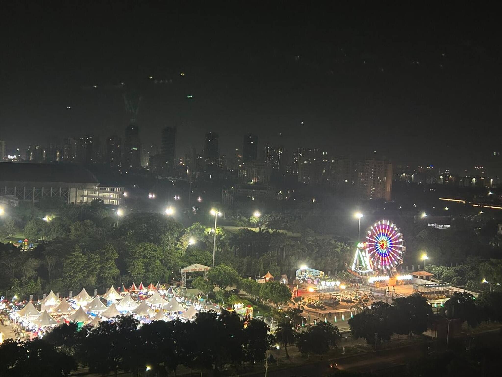 A cityscape at night with a prominent Ferris wheel and a densely packed area of tents.