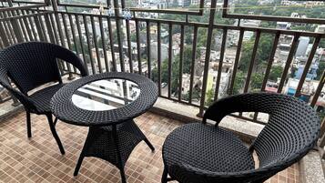 Een balkon met twee rieten stoelen en een glazen tafel, met uitzicht op een stadsgezicht met gebouwen en groen.