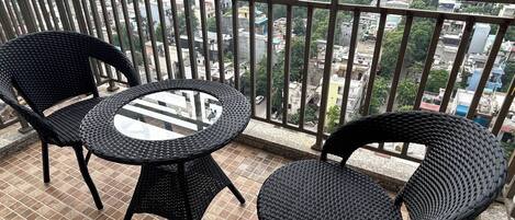 A balcony with two wicker chairs and a glass table, overlooking a cityscape with buildings and greenery.
