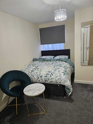 Ein Schlafzimmer mit einem Bett, einem Sessel, einem kleinen Tisch, einer Pendelleuchte und einem Fenster mit Jalousien.