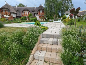 A residential area with a swimming pool, thatched-roof houses, and a paved pathway leading to the pool. - Spacious 3-bedroom apartment in tranquil Cserszegtomaj, Balaton view, pool, AC (Cserszegtomaj)
