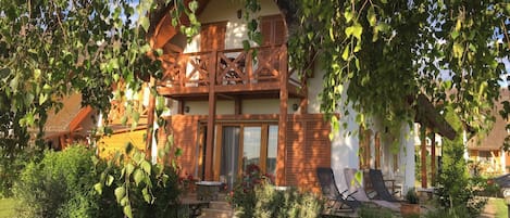 A two-story wooden house with a balcony, surrounded by greenery and a garden.