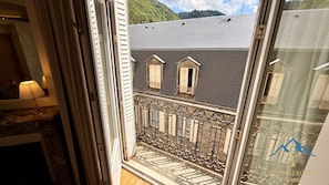 Ein Balkon mit schmiedeeisernem Geländer, ein Fenster mit Rollläden und Blick auf ein Gebäude mit grauem Dach.