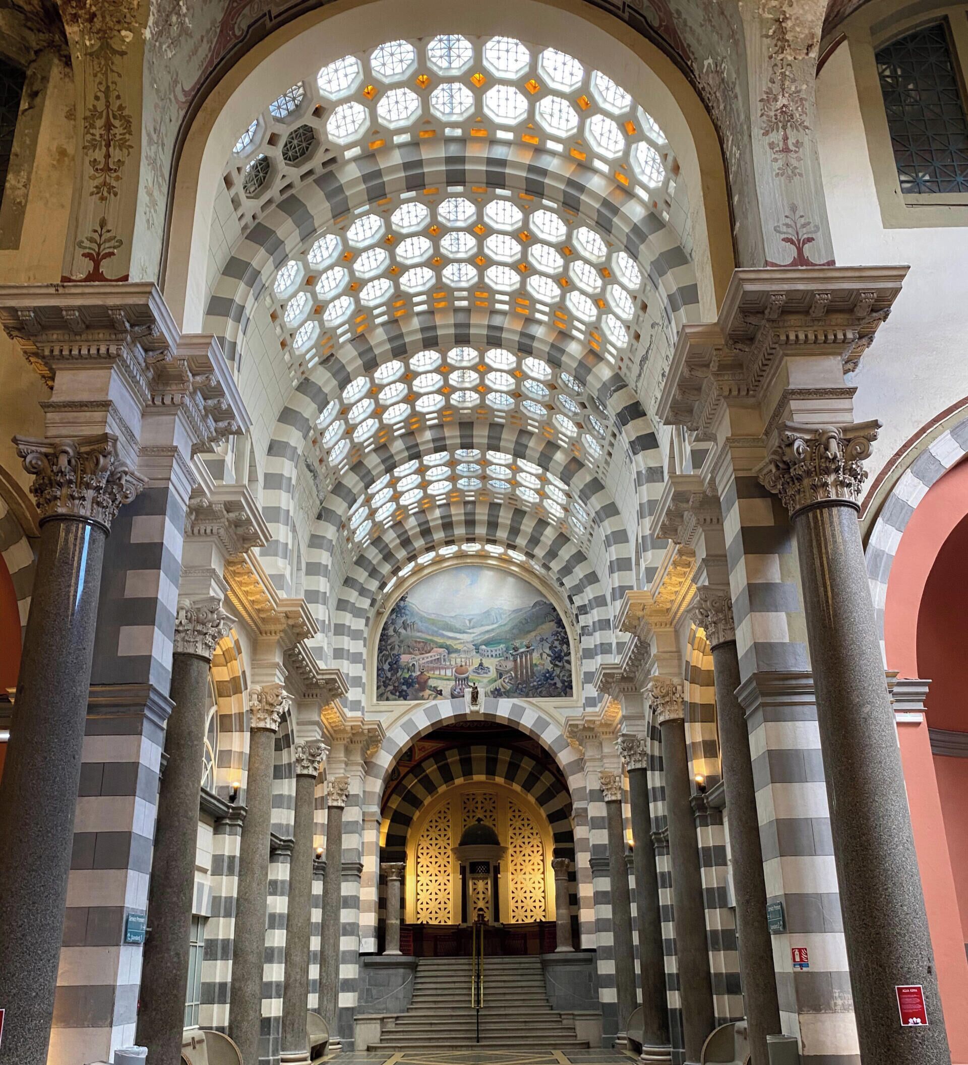 A grand hall with a high, arched ceiling featuring a geometric pattern of windows, flanked by tall columns with intricate capitals.