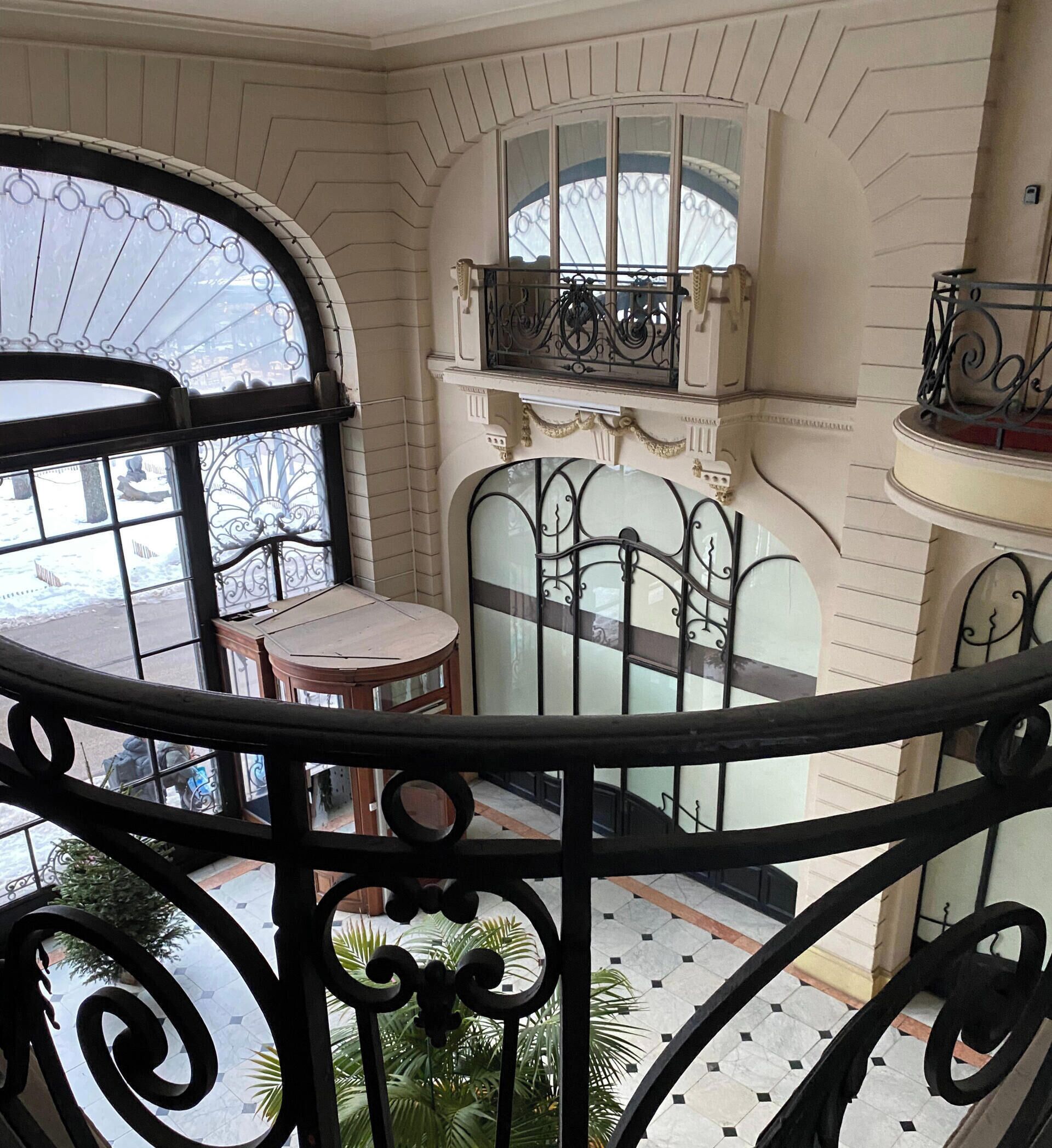 A balcony with a wrought iron railing, a small table, and a chair. There is a window with decorative ironwork and a view of the outside.