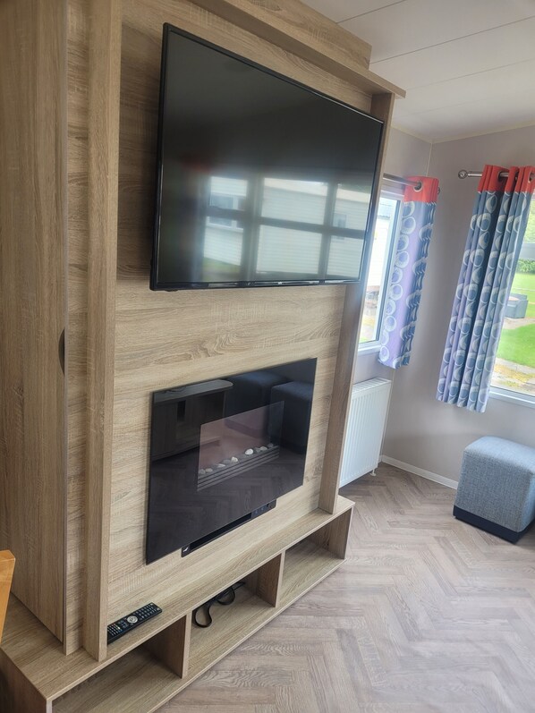 A modern living room with a flat-screen TV mounted on a wooden wall unit, a fireplace with a glass door, and a window with patterned curtains. - Beautiful Caravan minutes from Cartmel & Grange-over-Sands, Lakes are nearby. (Flookburgh)