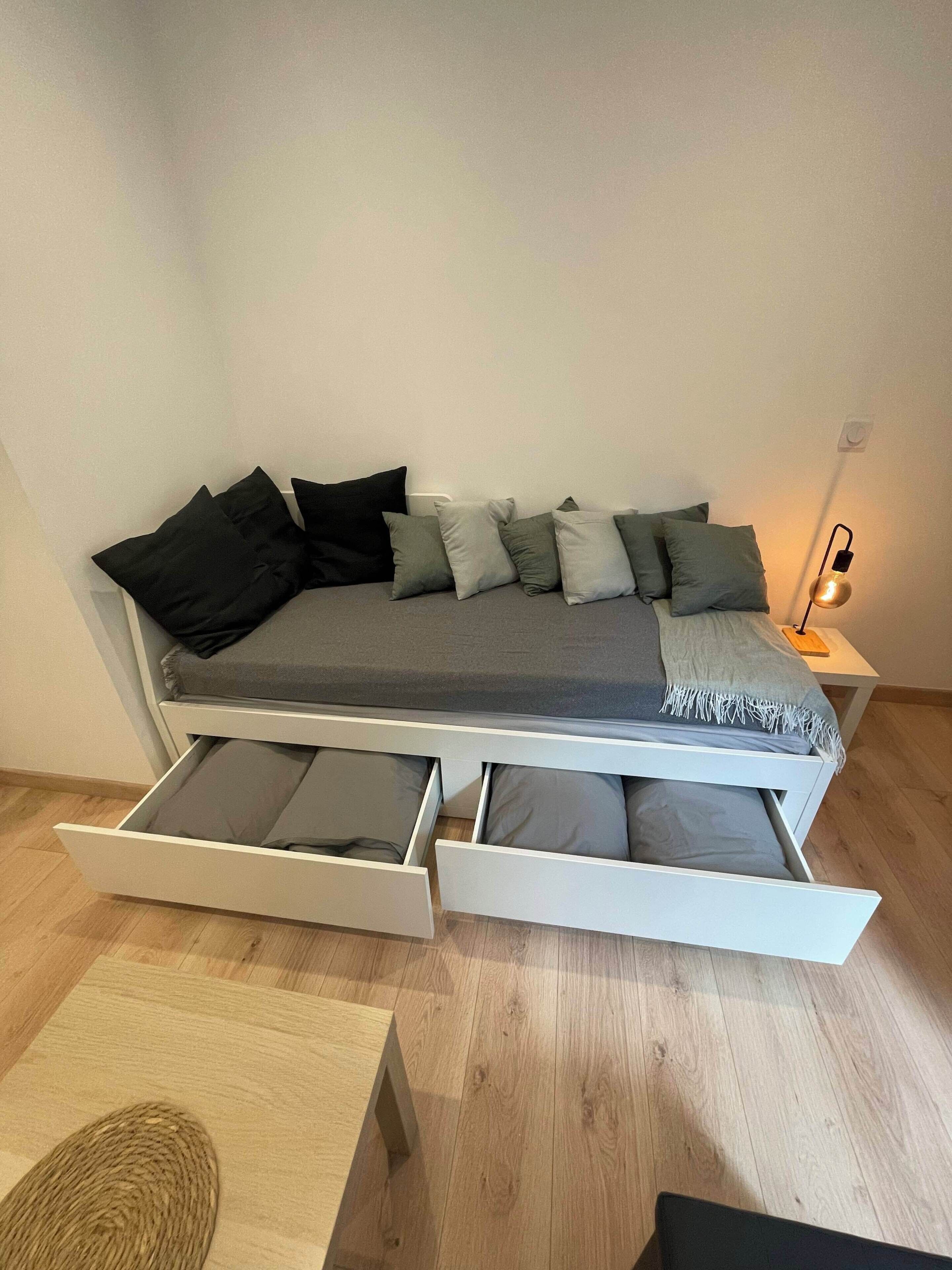 Een bed met opbergladen eronder, een nachtkastje met een lamp en een geweven vloerkleed.