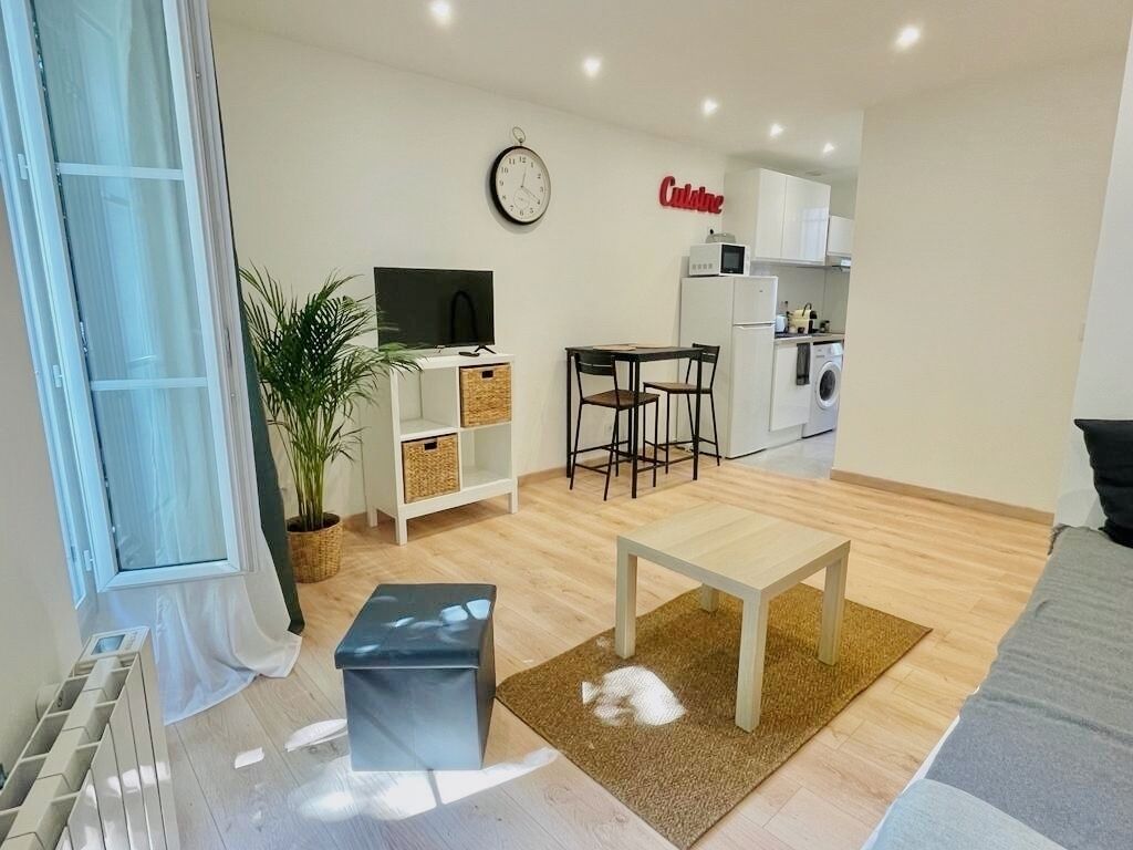 Una sala de estar moderna con un sofá, una mesa de centro, un televisor y una zona de cocina con refrigerador y lavadora.